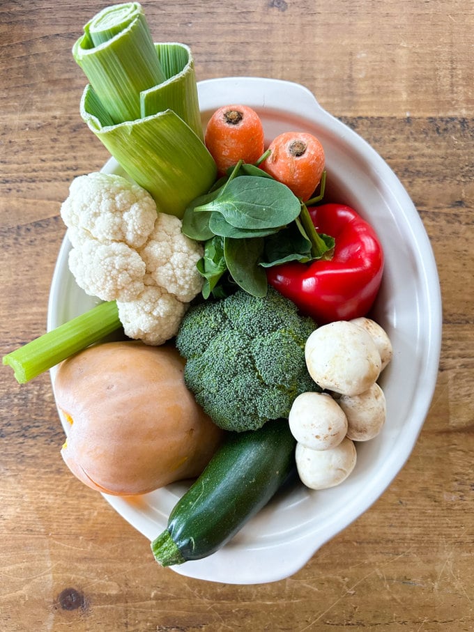 A white slow cooker packed with carrots, leek, cauliflower, celery, mushrooms, broccoli, red pepper and herbs.