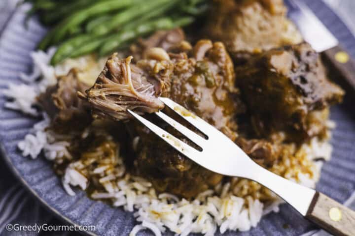 a forkful of tender meat with a plate of food with meat, rice and green beans in the background.