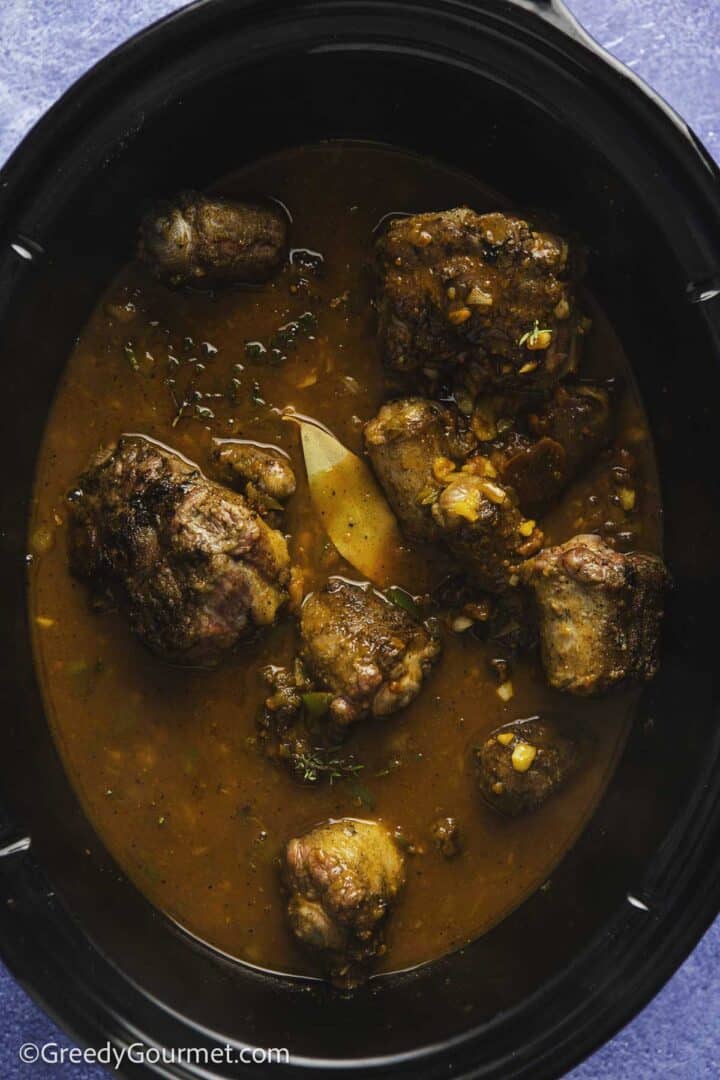 oxtail pieces doused in a reddish brown gravy with thyme sprigs and a bay leaf in a black pot on a blue background