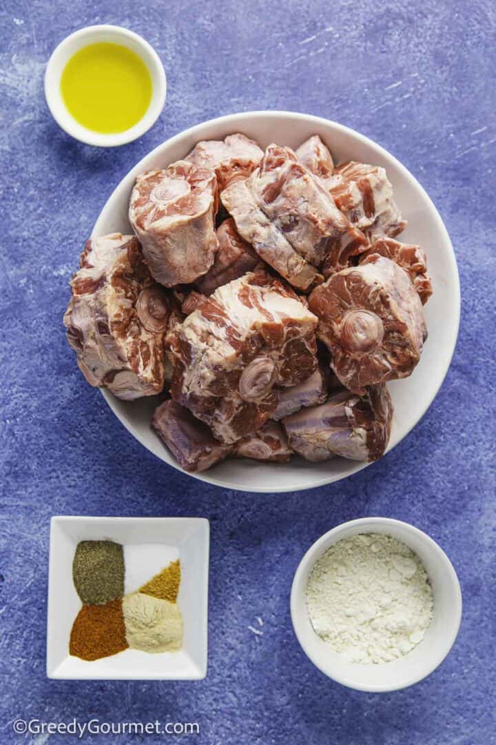 a bowlful of raw oxtail pieces, a small bowl of olive oil, another with flour, and another with spices, all placed on a blue background