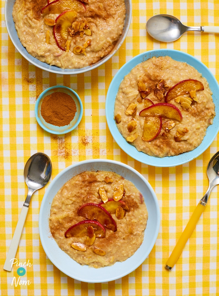 Pinch of Nom's Apple Pie Porridge served in three bowls on a yellow and white striped tabletop, with spoons waiting to dig in. Apple slices and flaked almonds are scattered on top.