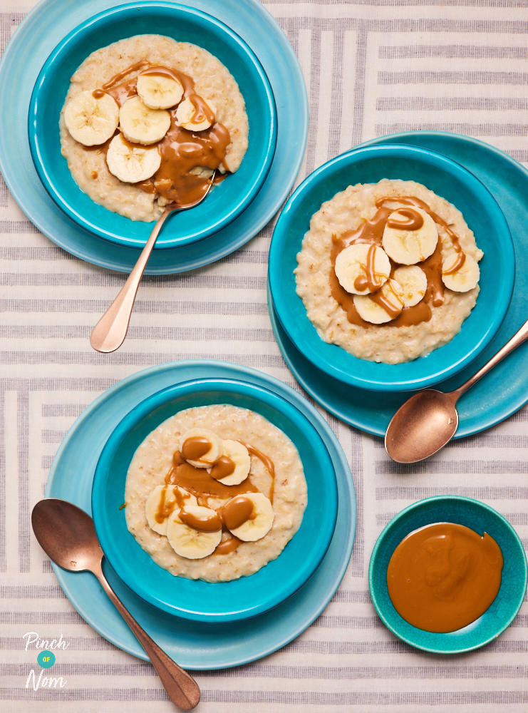 Three bowls of Pinch of Nom's Biscoff and Banana Porridge are served drizzled with melted Biscoff spread, topped with freshly-sliced bananas. A side bowl of melted Biscoff spread is on the side, with spoons waiting to tuck in.