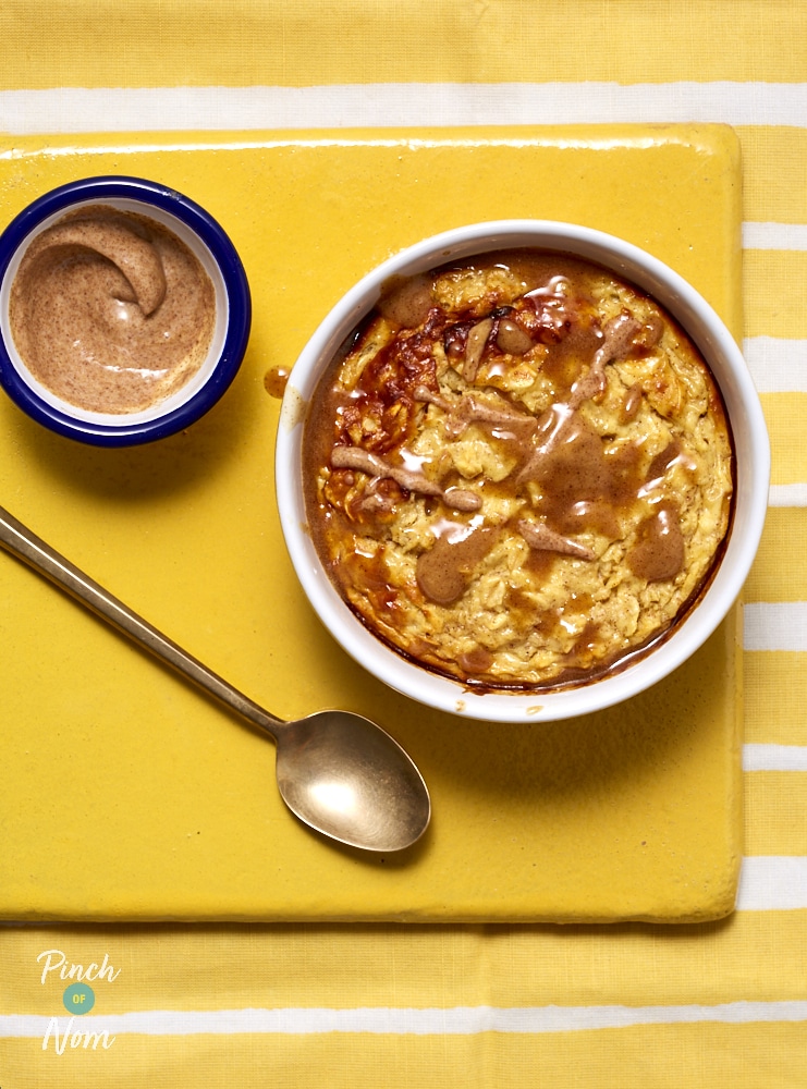 A bowl of Pinch of Nom's Cinnamon Roll Baked Oats is served on a bright yellow tabletop, with a pot of the sticky-sweet drizzle to the side, and a spoon waiting to tuck in.