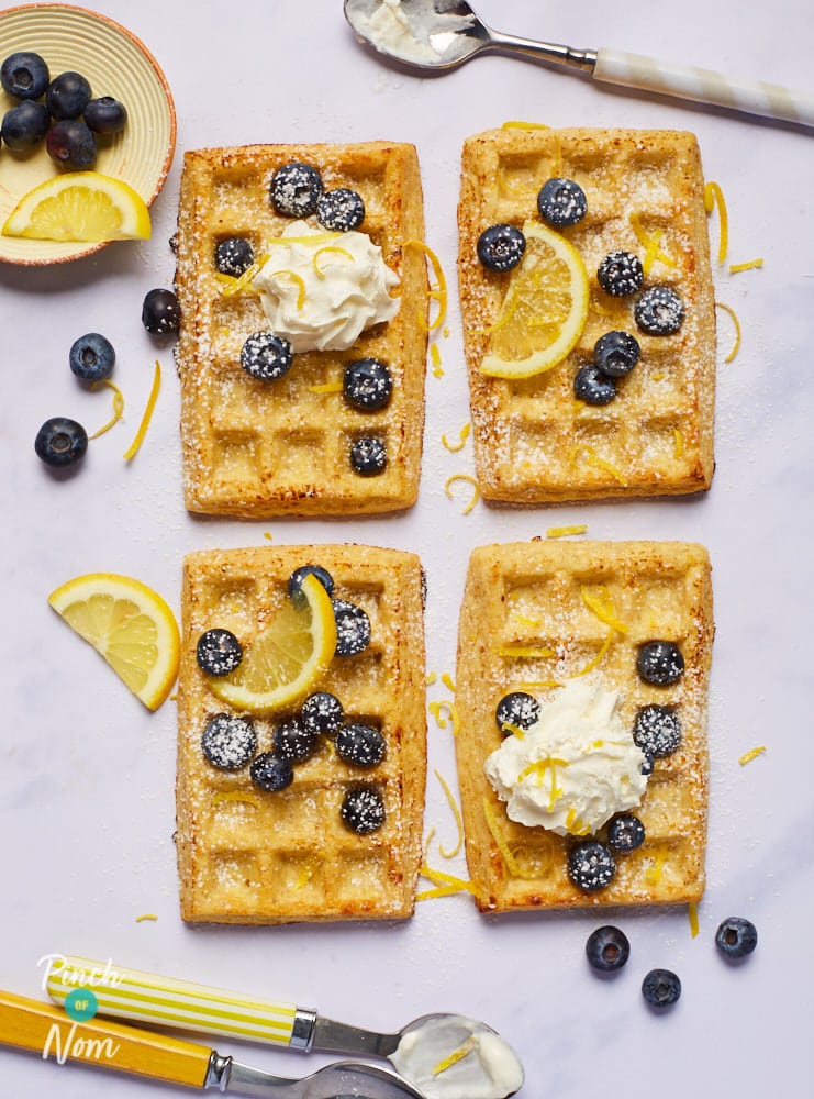 Four of Pinch of Nom's Lemon and Blueberry Waffles are served on a white tray with lemons, blueberries and a dollop of fat-free Greek yoghurt on top. There's a spoon to the side waiting to dig in.