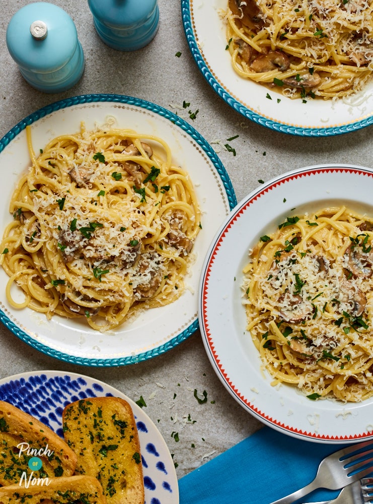 Three portions of Pinch of Nom's Mushroom Carbonara is plated up on a grey tabletop, with salt and pepper shakers at the centre. A portion of garlic bread is served to the side.