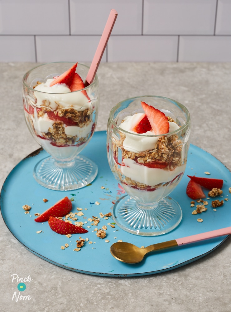 Two tall glass bowls are filled with Pinch of Nom's Peanut Butter and Jelly Pots. Yoghurt is layered with jam, strawberries and golden homemade granola, with spoons nearby, ready to dig in.