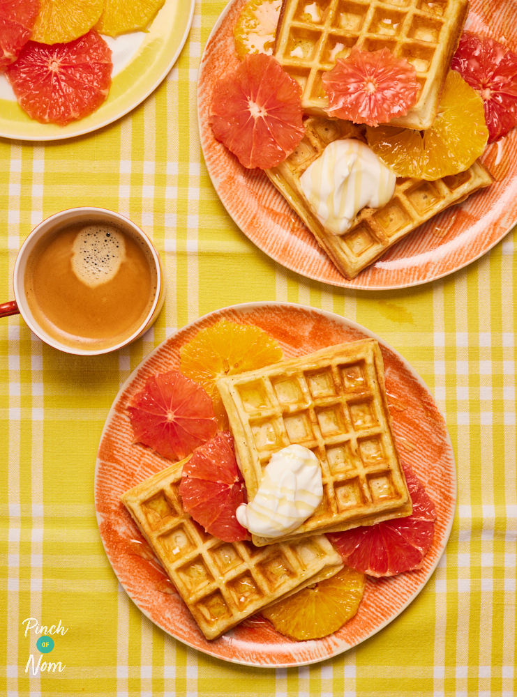 A table is set with two plates of Pinch of Nom's Sunshine Waffles, topped with chopped fruit and a dollop of yoghurt, paired with cups of tea.